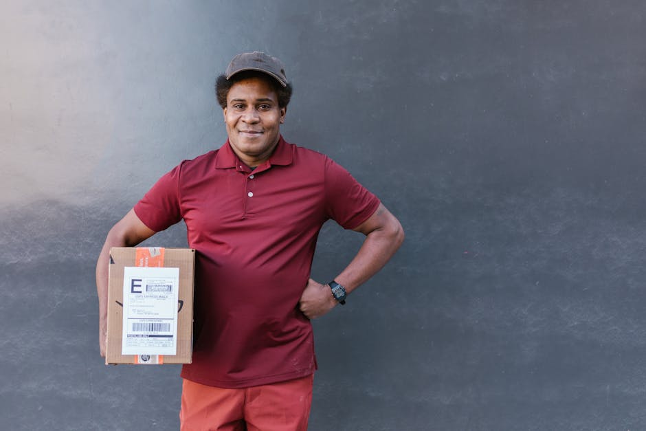 A smiling delivery person with medium brown skin and curly hair under a grey cap, standing against a plain dark grey wall. He is dressed in a maroon polo shirt and matching pants, holding a small card