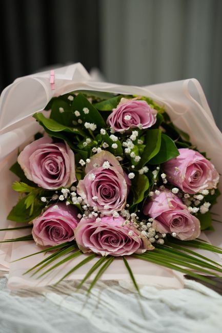 A bouquet of fresh, light pink roses with tightly curled petals is arranged with lush green foliage and small white baby's breath flowers, all wrapped in delicate white tissue paper. The bouquet is re