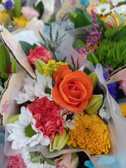 A close-up view of a vibrant floral bouquet featuring a large, peach-colored rose at the center, surrounded by white daisies, pink carnations, yellow chrysanthemums, and purple lavender sprigs. The fl
