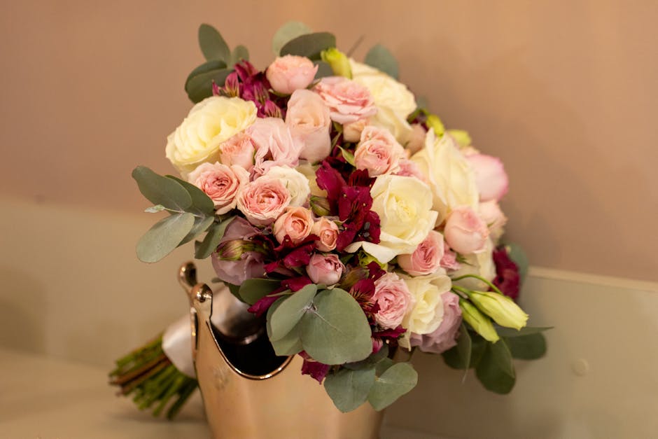 A bouquet arrangement of fresh flowers featuring pale pink roses, white roses, and deep burgundy flowers, accented with silvery-green eucalyptus leaves. The flowers are neatly clustered in a compact, 