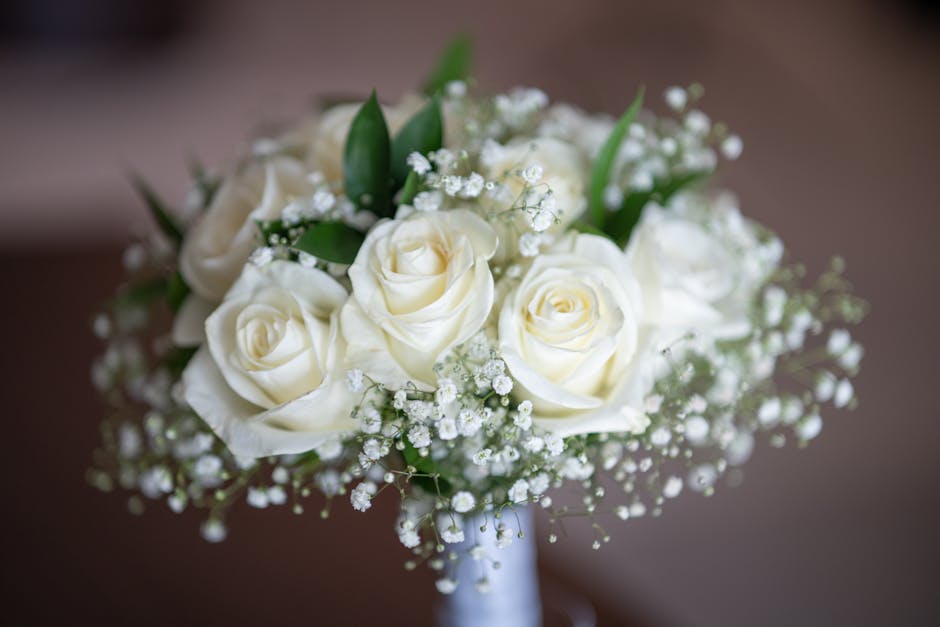 A bouquet arrangement consisting of creamy white roses with gently unfurling petals, accented by delicate sprigs of baby's breath with tiny white blossoms and lush green leaves. The flowers are tightl