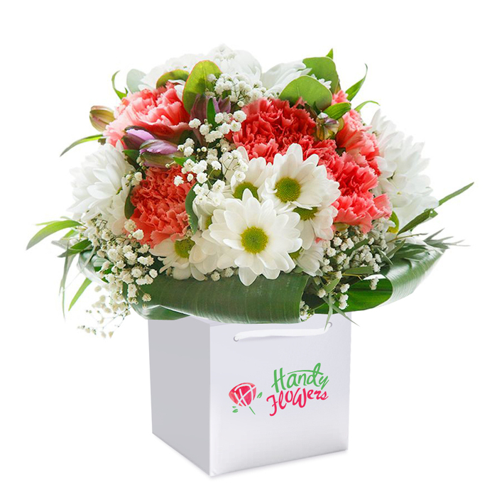 This beautifully arranged bouquet, standing proud in a crisp white square gift bag, captures the spirit of Bexley with meticulous artistry and fresh, local flair. A luxurious blend of soft coral and deep red carnations, each bloom bursting with textured, frilled petals, takes centre stage and instantly draws the eye-echoing the vibrant colours of spring gardens found in Old Bexley's leafy enclaves. Interspersed amid the carnations are pure white daisies, each with an inviting lime-green center that offers an elegant balance and cheerful pop of colour akin to wildflowers along the banks of Danson Lake. Fine sprays of white baby's breath scatter delicately throughout, lending an airy texture that lightens the bouquet and brings to mind the freshness of early mornings on Bexley's village greens. Glossy, deep green leaves form a lush, enveloping collar around the base, providing both support and visual harmony, with a few gently curved, unopened buds tinged with subtle hues of purple and soft pink peeking out for added depth and promise. The bouquet's rounded, full shape radiates a sense of abundance and presents an inviting invitation for any celebration, be it a milestone anniversary in Blendon or a heartfelt birthday surprise along the Broadway. The bag's simple satin handles suggest ease of transport, making it ideal for same-day delivery across Bexley and surrounding neighbourhoods. The detailed, polished presentation expresses the freshness, quality, and care that Bexley Flowers is renowned for, making this arrangement a perfect choice for marking life's special occasions or simply conveying a generous, joyful sentiment.