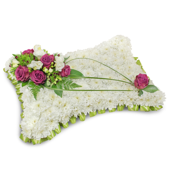 A traditionally elegant sympathy pillow presented on a clean white field, its gentle shadow grounding the arrangement and underscoring the satin sheen of the border. The pillow's surface is densely covered with hundreds of small, snow-white chrysanthemum florets; their tightly packed petals form a soft, cushion-like texture that conveys tranquility and purity. At the top-left a vivid cluster of deep magenta roses provides a poignant focal point-the roses' velvety petals unfolding with quiet dignity-accompanied by delicate white freesia or lisianthus that introduce a light, floral perfume and a fragile contrast. Rich green foliage, from lacy fern fronds to broader glossy leaves, frames the accent cluster and enriches the composition with natural depth. Three long, arching stems of bright bear grass sweep across the white surface in an artful curve, guiding the eye toward the lower-right where a solitary magenta rose echoes the main grouping and balances the design. A neat trim of folded lime-green satin ribbon encircles the pillow, forming a soft ruffled edge that both defines and softens the shape. The lighting is soft and even, accentuating the tactile interplay between the smooth satin, the velvet of roses and the feathery mums. Prepared with the respectful care of a local florist, this arrangement-available from Bexley Flowers (bexleyflowers.org.uk) for memorial occasions around Danson Park, Bexleyheath and nearby villages-serves as a lasting expression of condolence and peaceful remembrance.