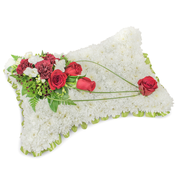 A serene funeral cushion, sculpted into a soft rectangle and photographed to show every floral texture, begins with an immaculate base of densely packed white chrysanthemums that blanket the form like freshly fallen snow. The tiny petals create a plush, tactile surface that suggests comfort and repose, the kind of quiet presence meant to console. A narrow lime-green border of satin ribbon or tender foliage edges the pillow, giving a whisper of colour and a clean silhouette. At the upper left, a richly hued cluster of deep red roses and dark burgundy carnations forms a focal point; their velvety petals and ruffled layers contrast with the white backdrop, while delicate sprays of white freesias and small spray chrysanthemums add soft highlights and a faint, sweet fragrance. Lush green leaves, including feathery fern fronds, sit behind the blooms, adding depth, structure, and an earthy freshness. From the main spray a trio of slender, bright green stems - likely bear-grass or decorative foliage - describes an elegant arc across the chrysanthemum surface toward the lower right, where a single perfect red rose rests, completing the visual sentence and symbolising steadfast love and remembrance. The lighting is gentle, natural-looking, emphasizing texture and shadow rather than gloss, and the overall impression is one of dignified tenderness. This timeless sympathy design is the kind of memorial piece often requested by families across Bexley, from Bexley Village to Barnehurst, for funeral chapels, wakes, and graveside ceremonies.